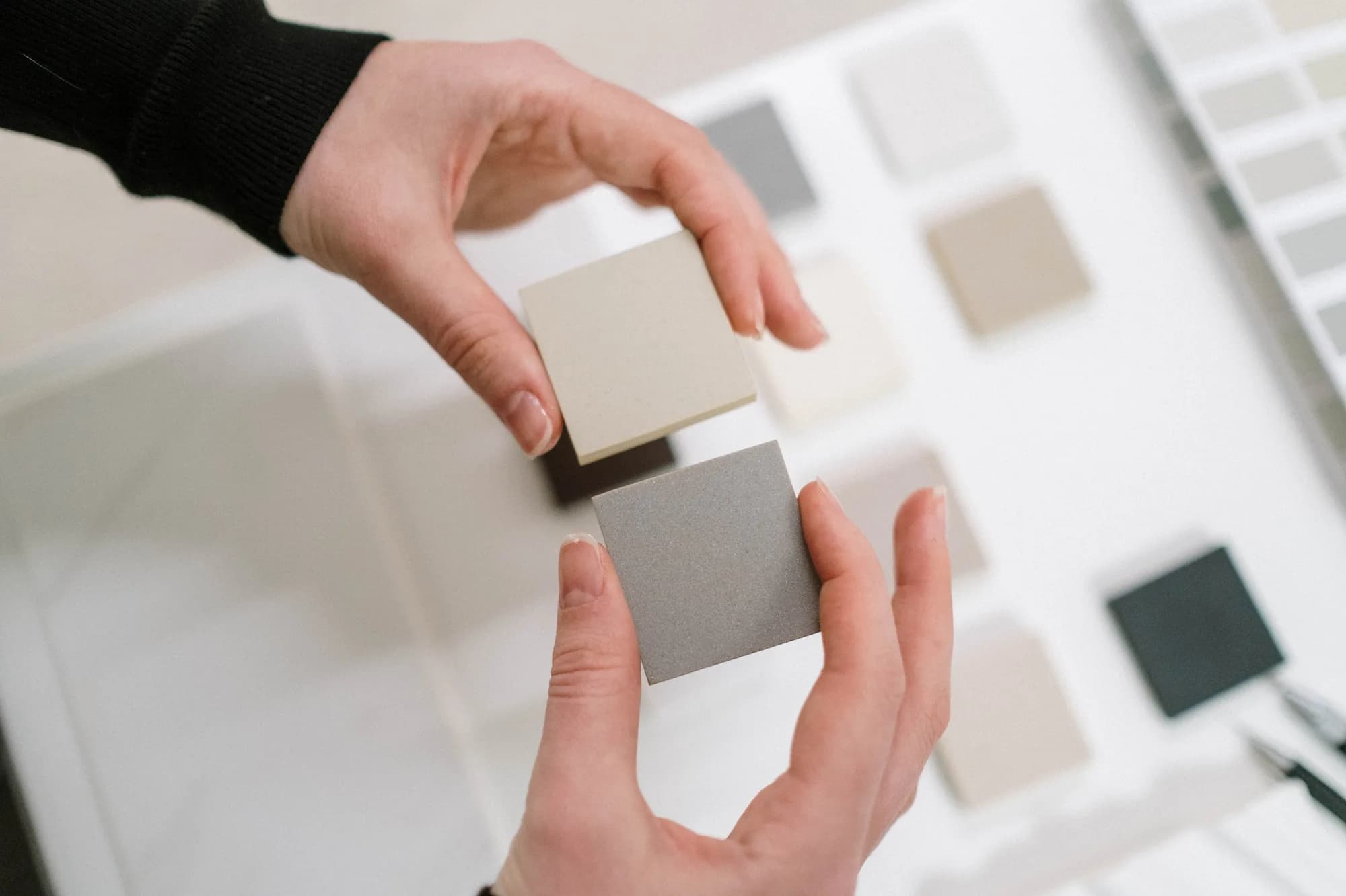 Deux mains comparent un échantillon crème et un échantillon gris au-dessus d’une table couverte d’échantillons de matériaux.
