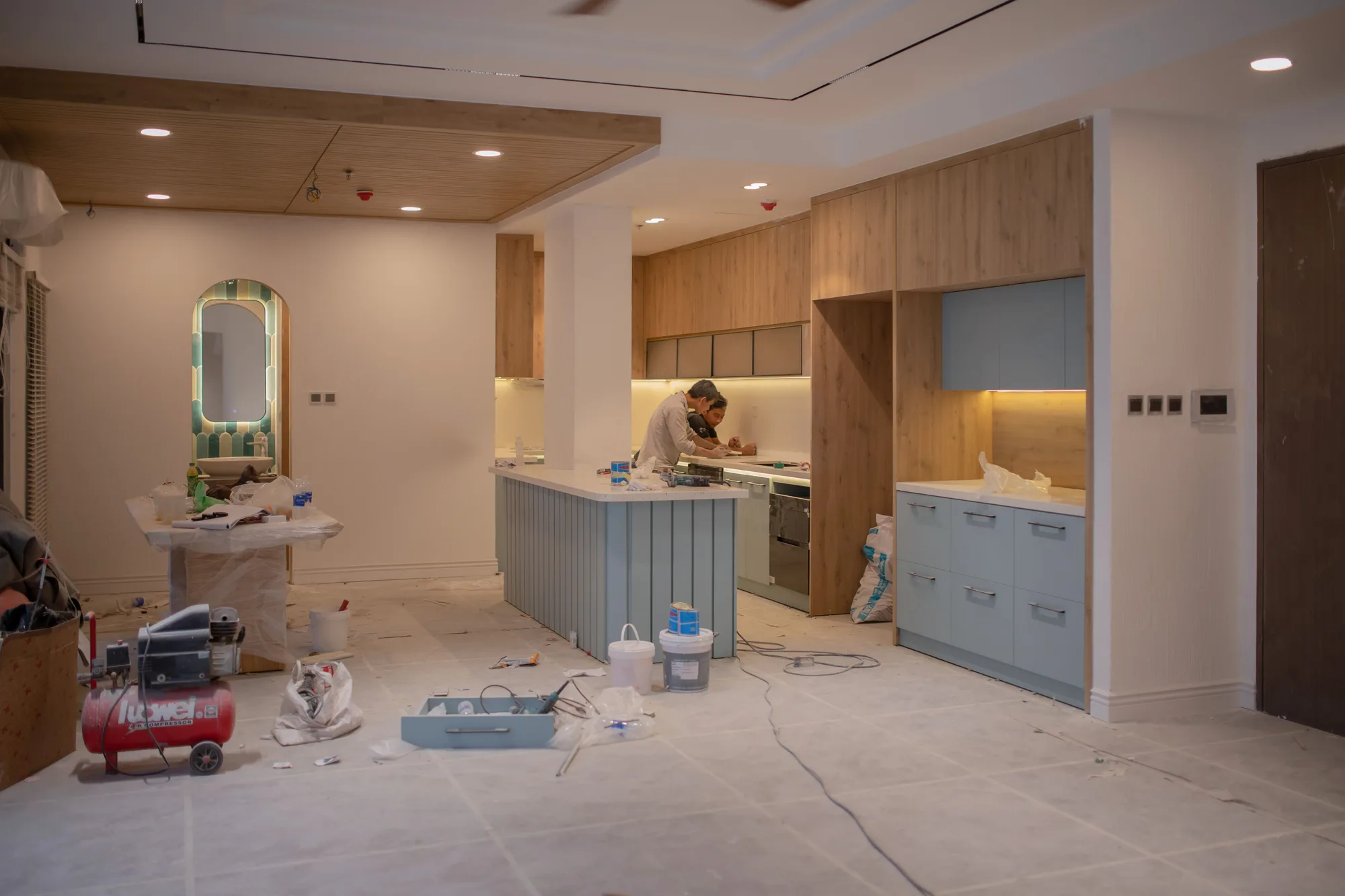 Cuisine en cours de rénovation : deux artisans au travail sur l’îlot, meubles en bois et bleu clair, sol protégé, compresseur et outils au premier plan.