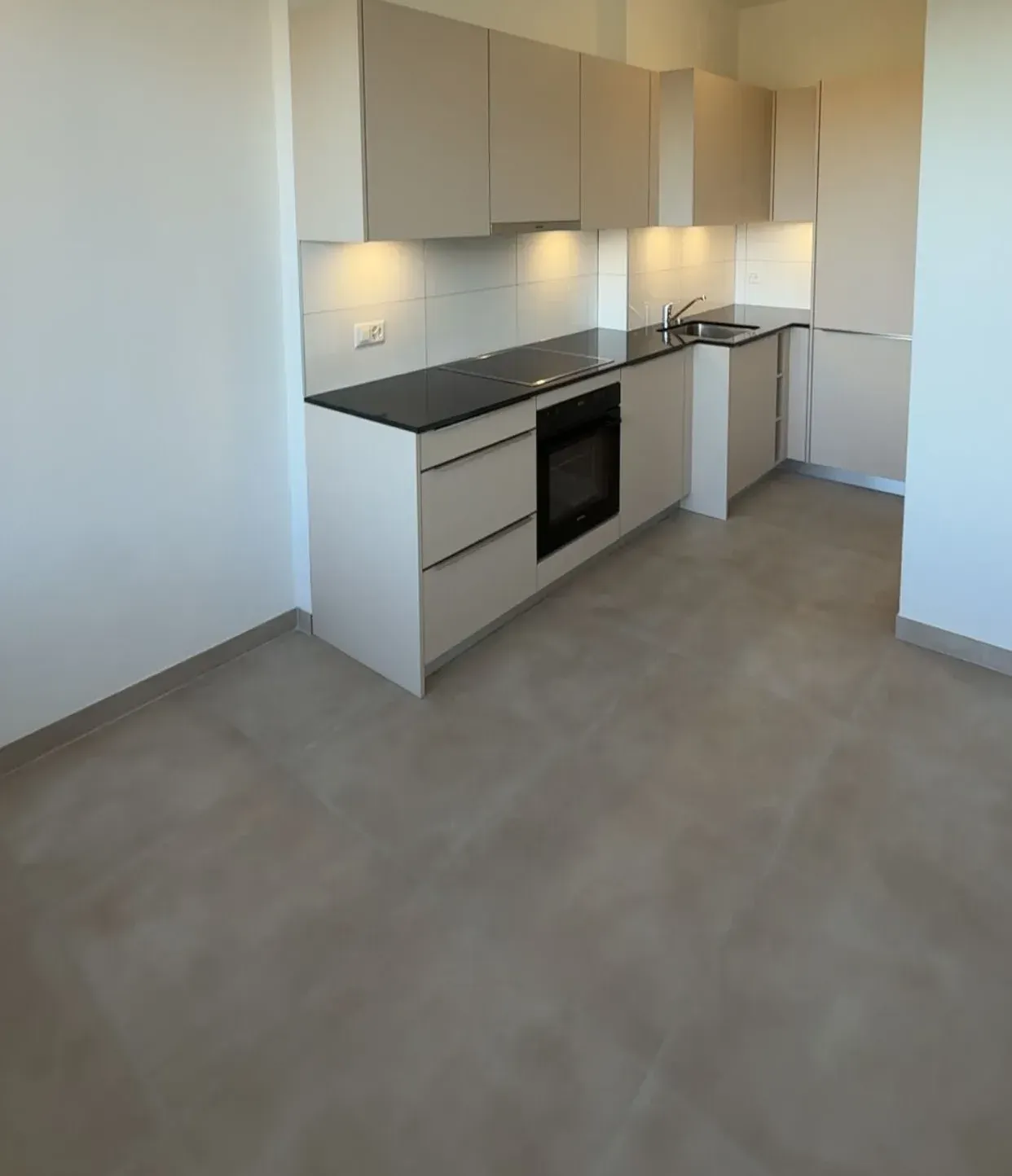 Cuisine en L aux façades beige taupe, plan de travail noir, crédence en grands carreaux blancs éclairée par-dessous et sol en grands carreaux gris beige.