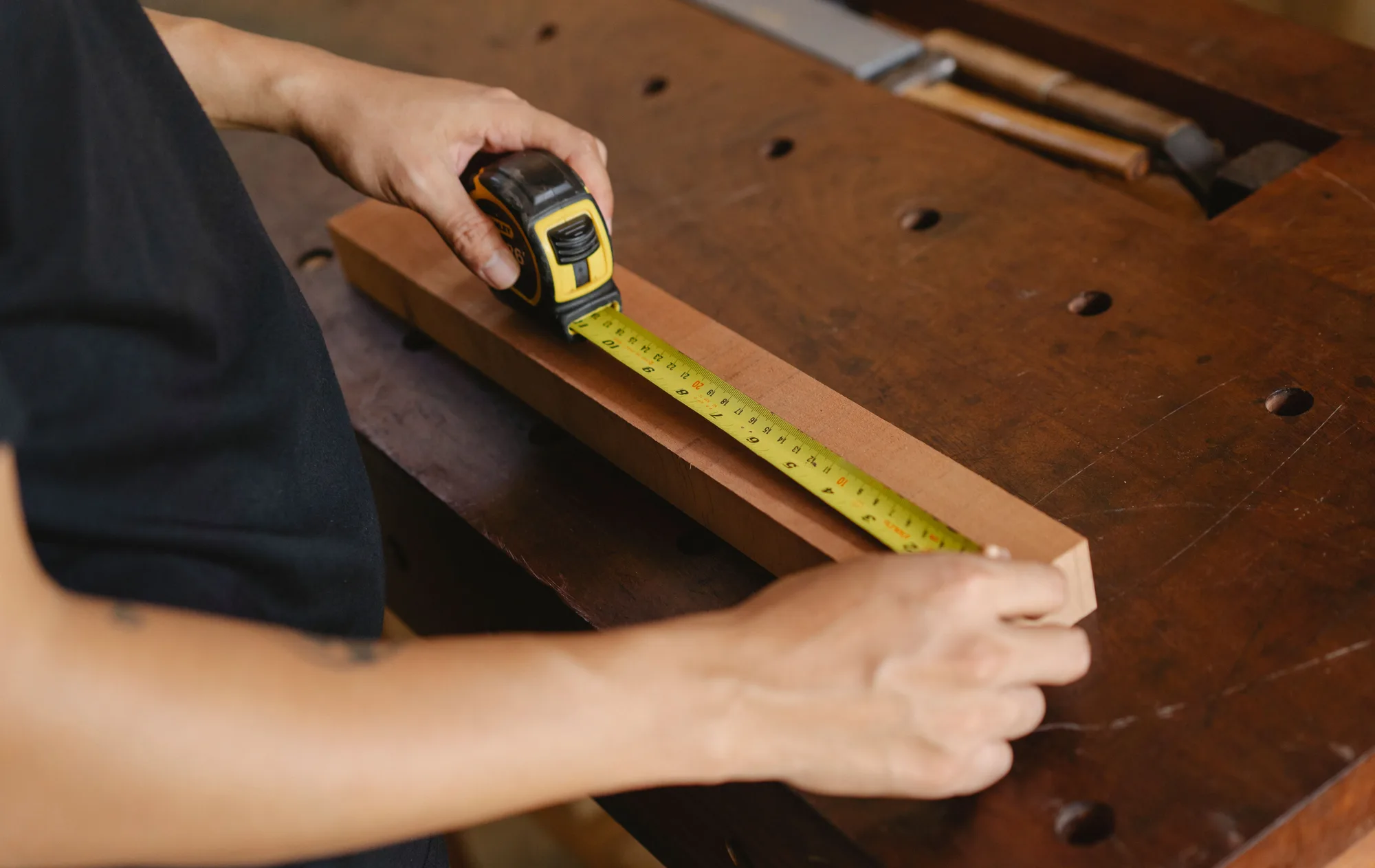 Mains qui mesurent une planche de bois au mètre ruban sur un établi en bois sombre.
